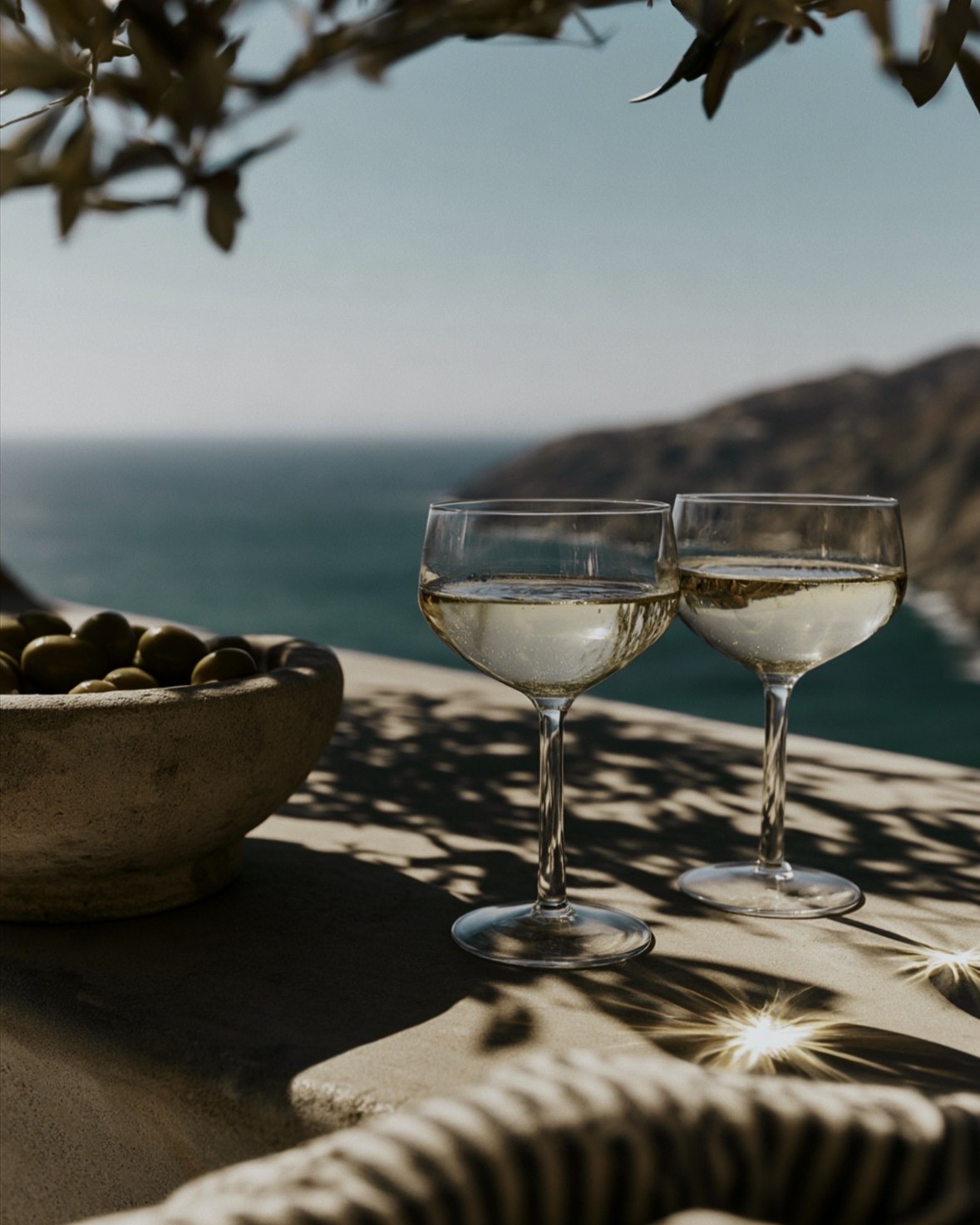 Two coupe glasses and a bowl of olives on a stone ledge above the sea
