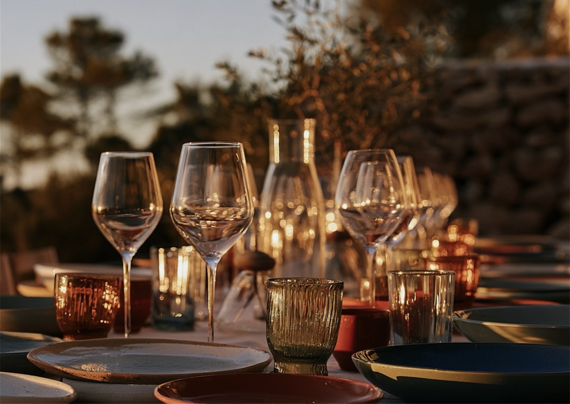 A long table laid outdoors at dusk, wine glasses and candle holders catching the last light