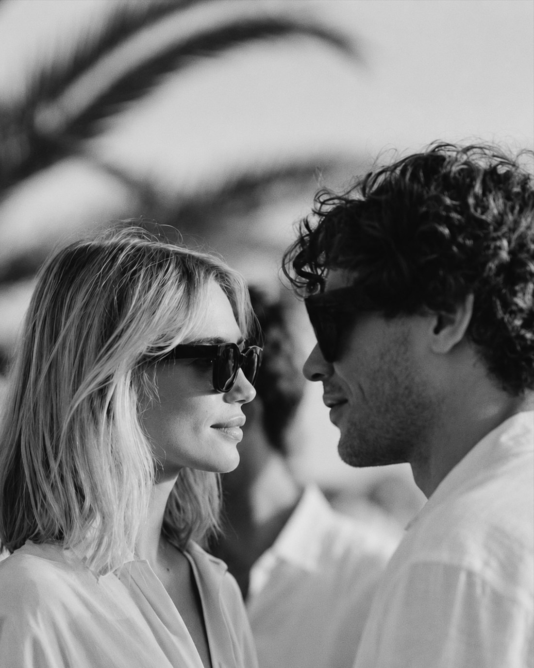 A woman and a man in sunglasses smiling at each other in conversation, palm fronds behind them